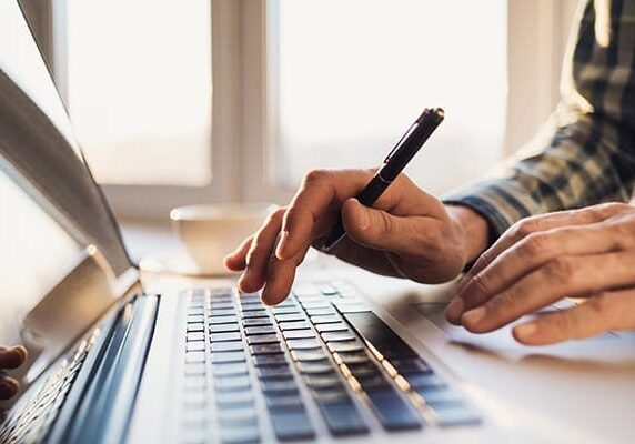 person holding pen and typing on a laptop