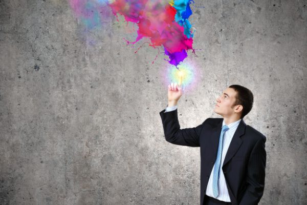 man in suit in front of concrete wall holding finger up which is alight and emitting splattered paint in many colors above it