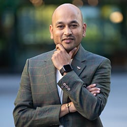 Ajit Nawalkha headshot photo, smiling man wearing a suit with one hand on arm and the other on his chin outside