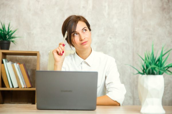 woman thinking with pen resting on head sitting in front of a laptop at a table in a home office