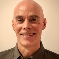 Charles Bassard headshot photo, smiling man with dark collared shirt in front of light background