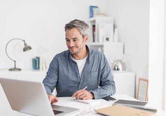 man working in an office at a white desk with his laptop and notebook