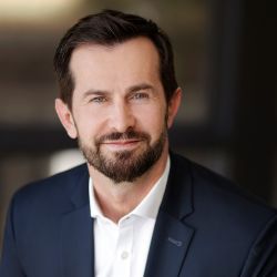 Slaven Drinovac PCC headshot photo, man with dark hair and beard in a dark blazer and white shirt smiling, blurred background