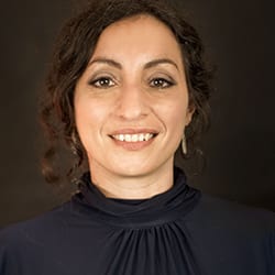 Erica Sosna headshot photo, smiling woman with dark hair wearing a black top in front of a dark brown background