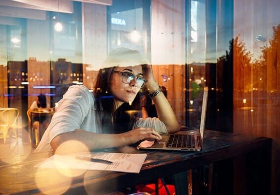 woman sitting at coffee shop window hightop working on her laptop at dusk