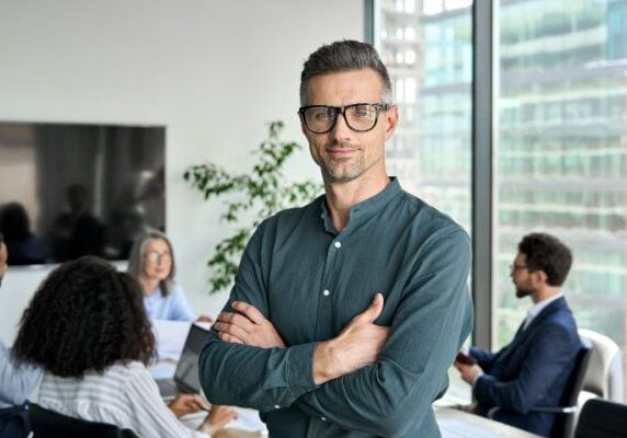 confident man smiling while others meet at a conference table behind him