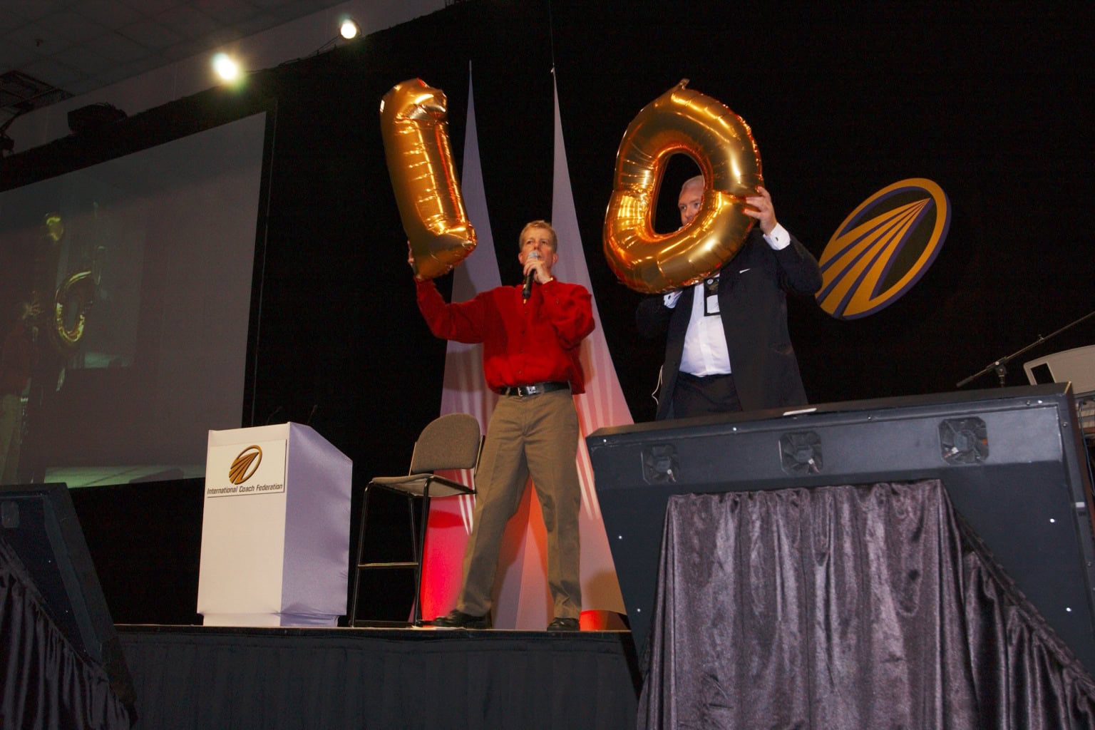 Two men stand on stage holding gold balloons—one displaying the number "1" and the other displaying the number "0"—celebrating the 10th anniversary of the International Coaching Federation at the annual conference.