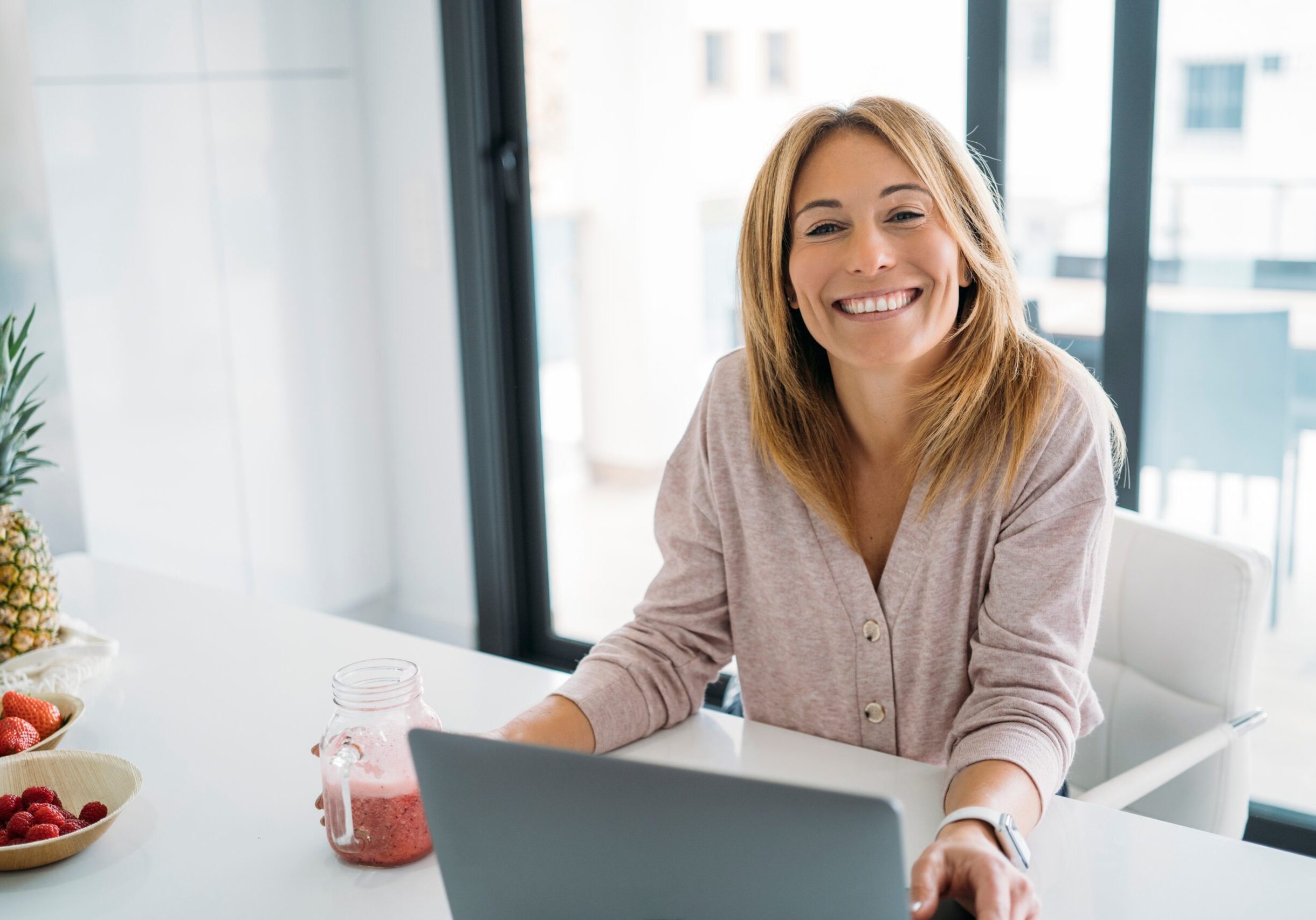 Woman smiling while working on a laptop at a kitchen table with a smoothie