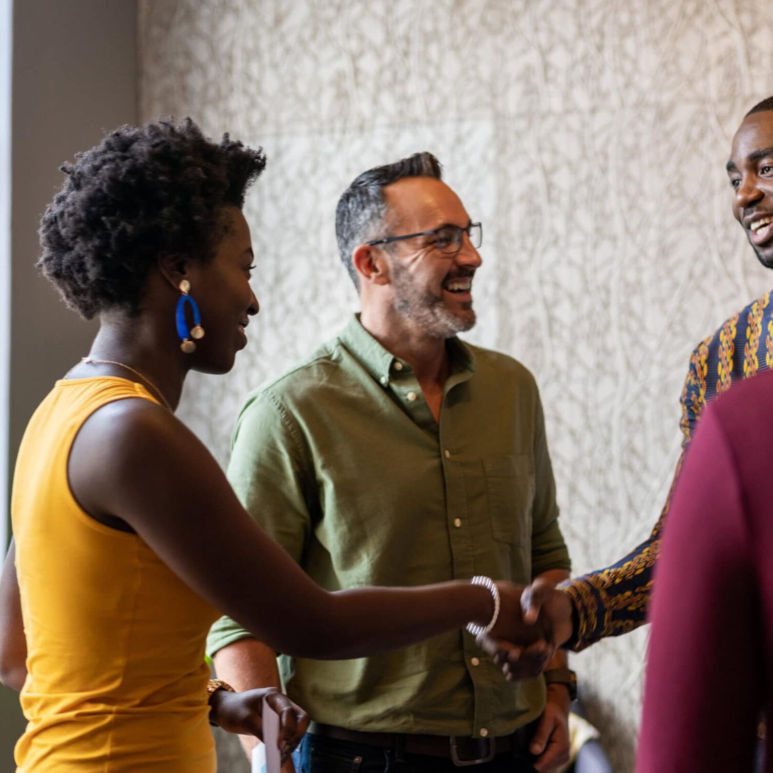 A diverse group of coaches connect warmly, shaking hands and smiling in an office setting. Two individuals greet each other in the foreground, while others stand nearby, fostering a friendly and collaborative atmosphere.