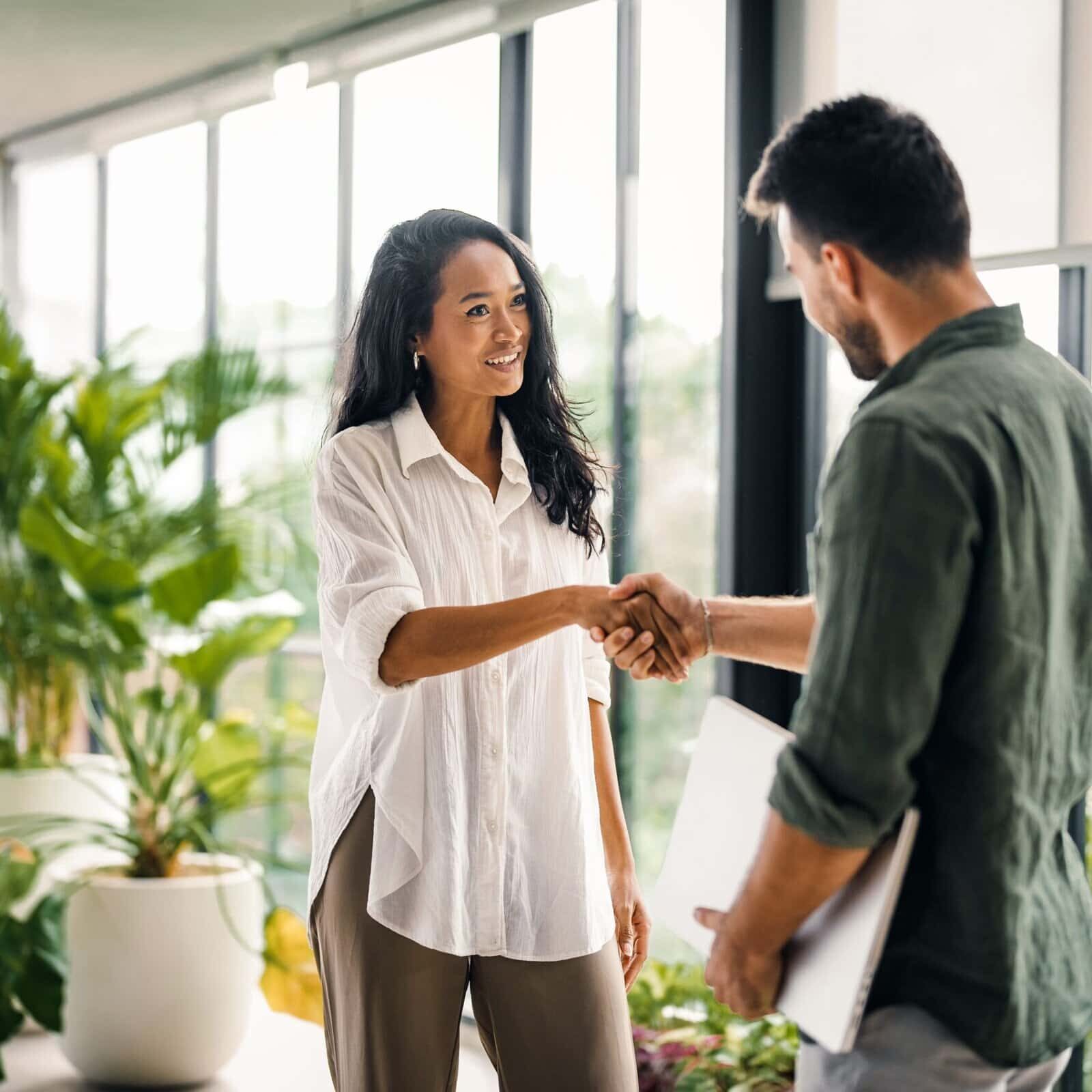 Two professionals shaking hands in a modern office with large windows and indoor plants.
