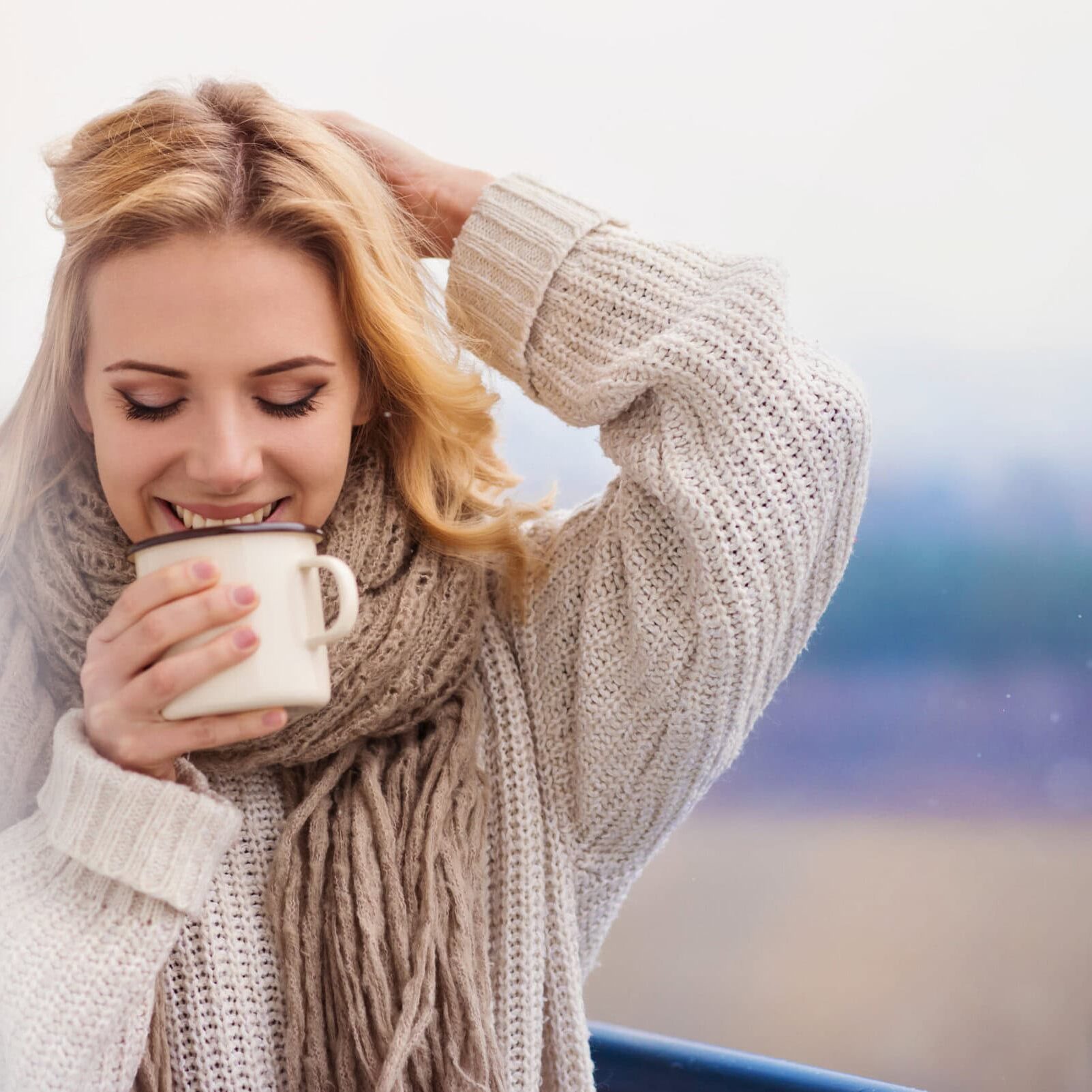 Woman in a knit sweater smiling while holding a mug outdoors on a balcony.