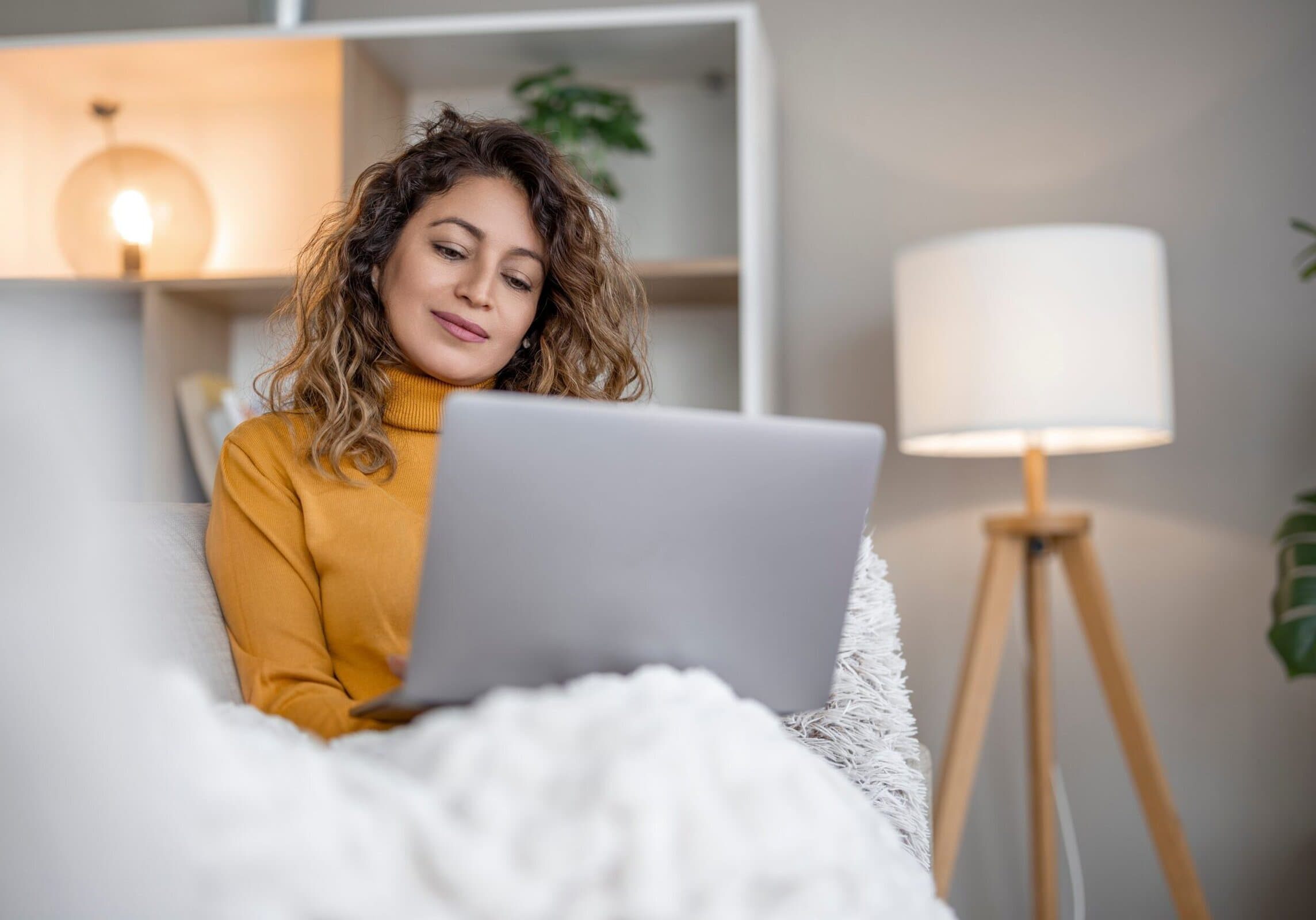 A woman with curly hair sitting on a couch at home while using a laptop in a softly lit living room.