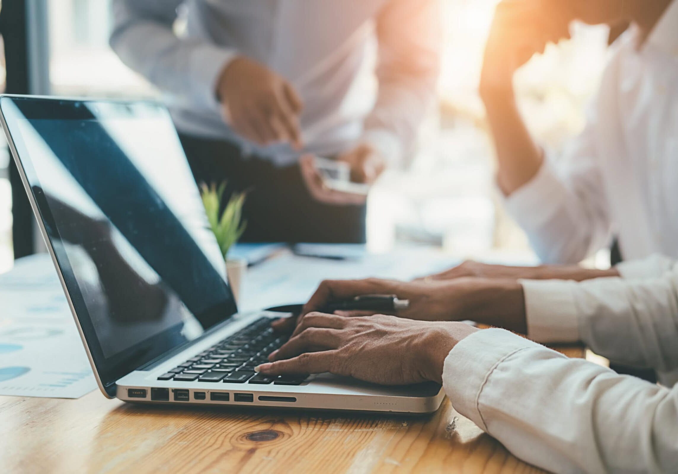 Hands typing on laptop while colleagues discuss ideas in office