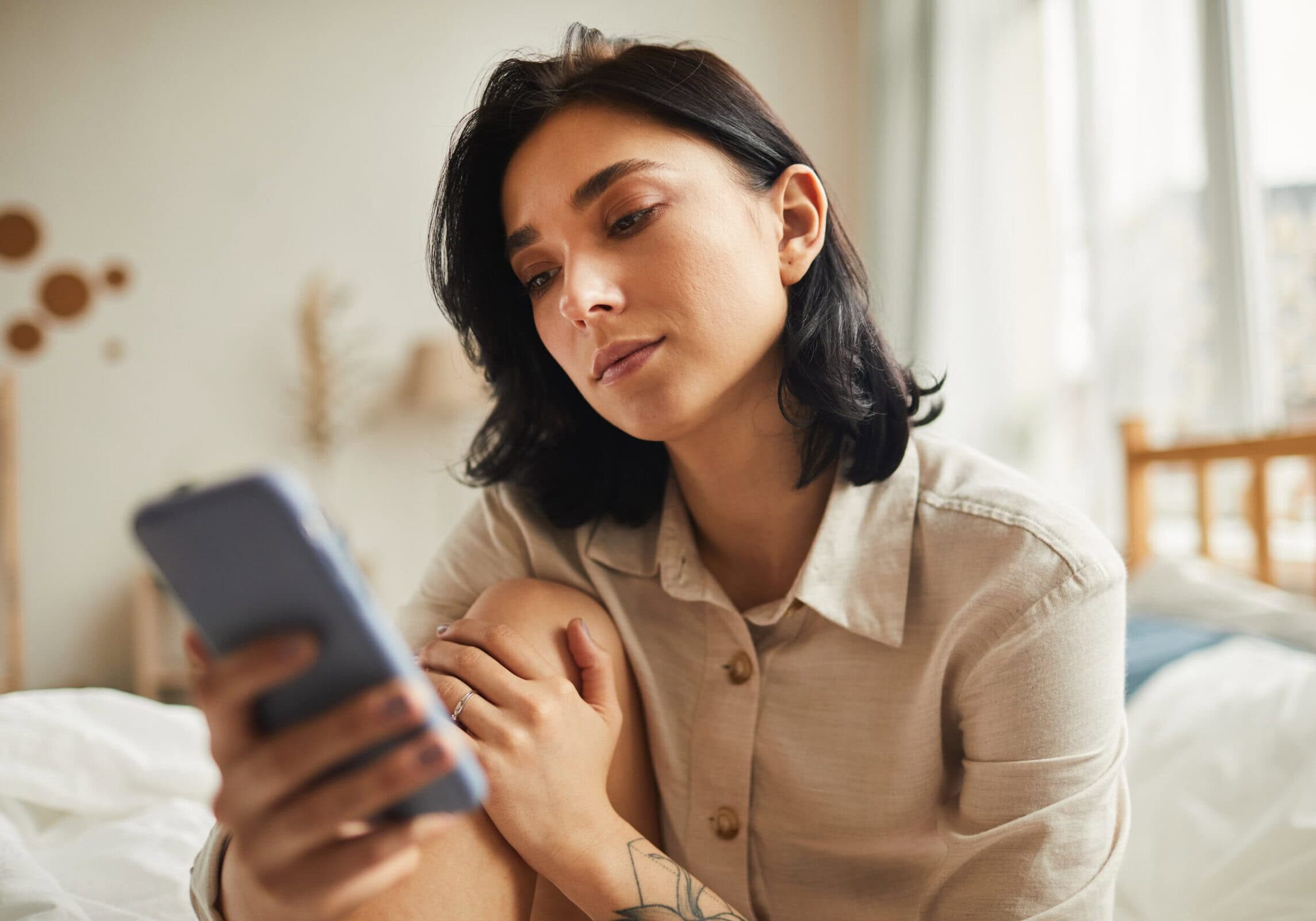 Woman sitting on bed looking at smartphone in softly lit room