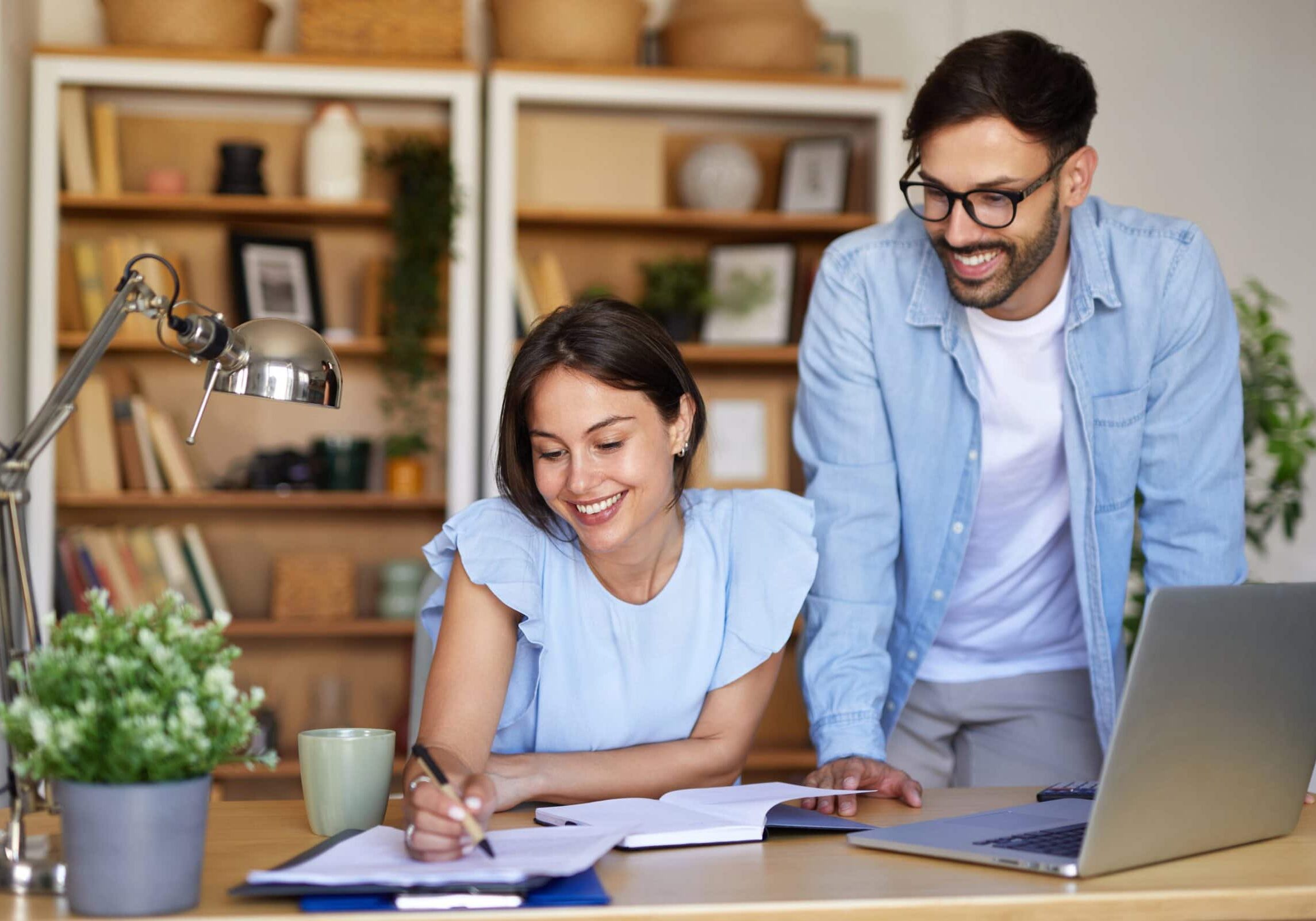 Two professionals working together at a home desk with laptop and notebook.