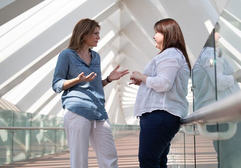 Two women engaged in a professional coaching conversation in a bright, modern architectural hallway.