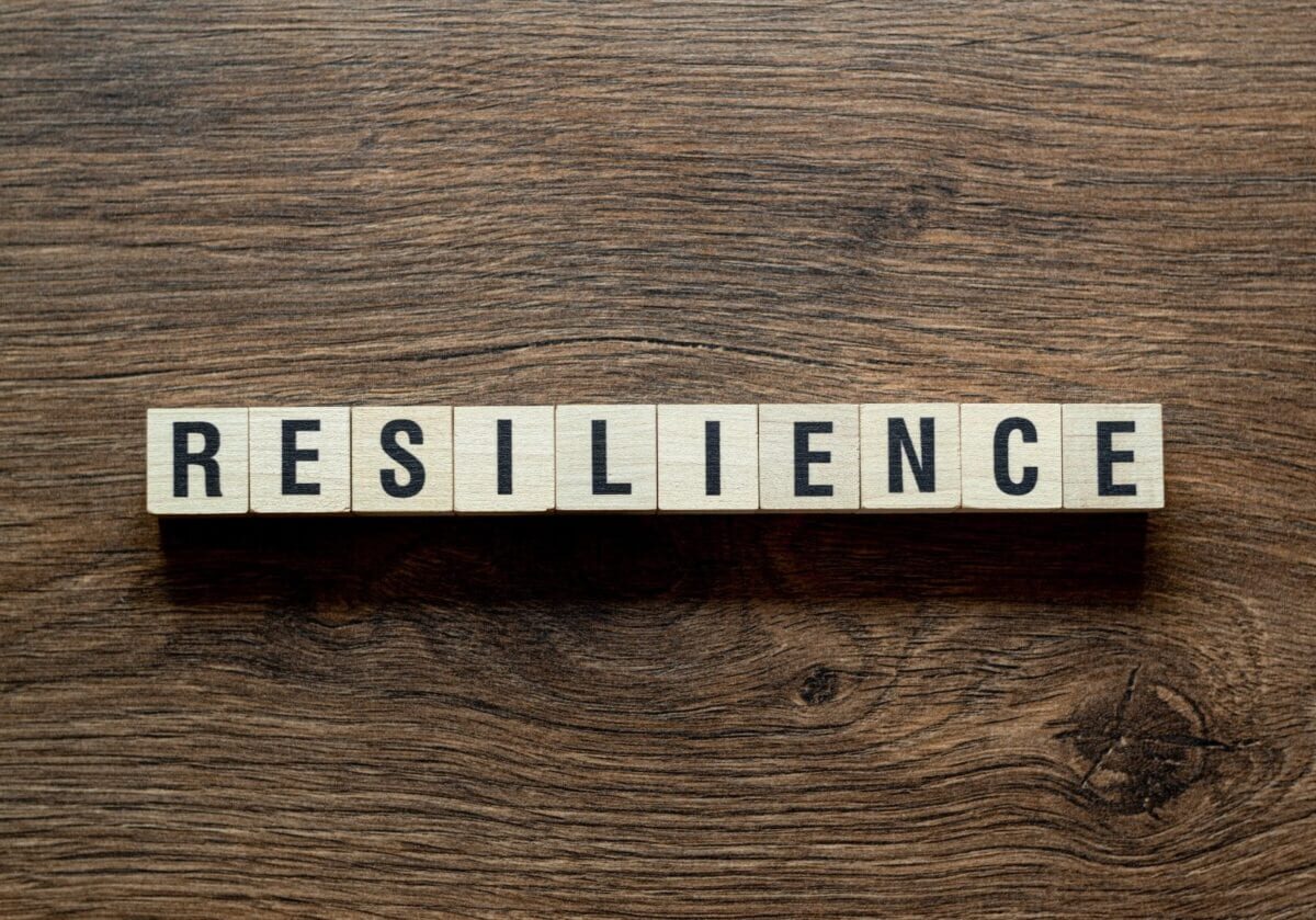 he word RESILIENCE spelled out in wooden letter tiles on a dark wood grain surface.