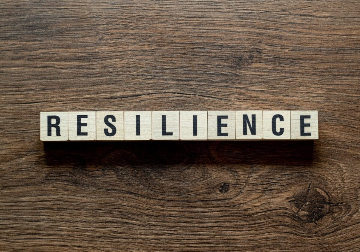 he word RESILIENCE spelled out in wooden letter tiles on a dark wood grain surface.
