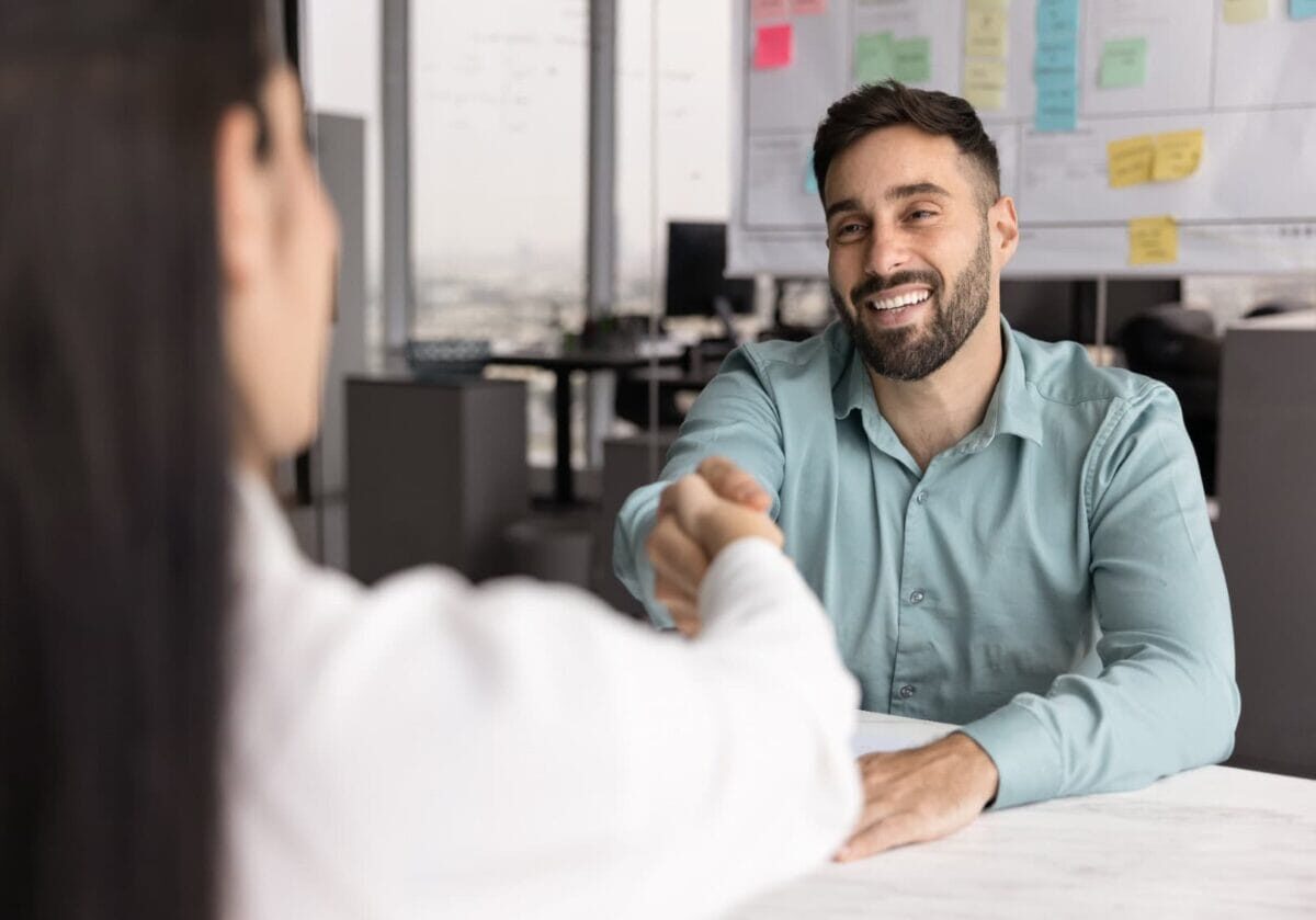 A man and a woman shake hands across a desk in a modern office.