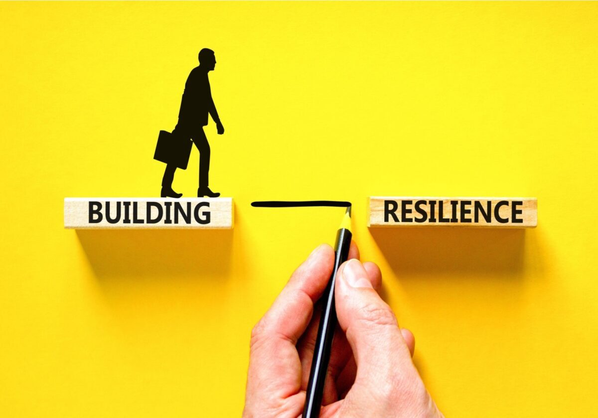 A hand draws a bridge between two blocks labeled "Building" and "Resilience" as a silhouette of a person walks across, symbolizing growth through challenge.