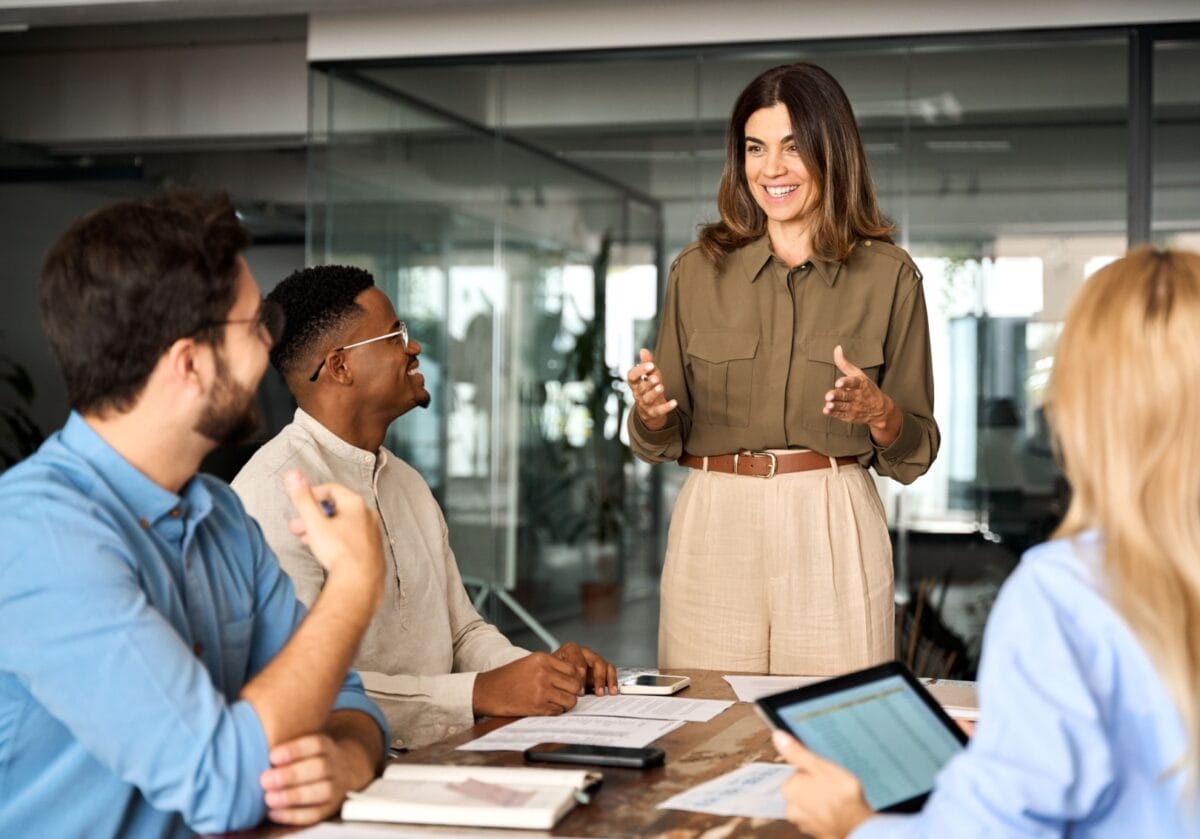 Smiling woman leading a meeting with three seated colleagues in a modern office.
