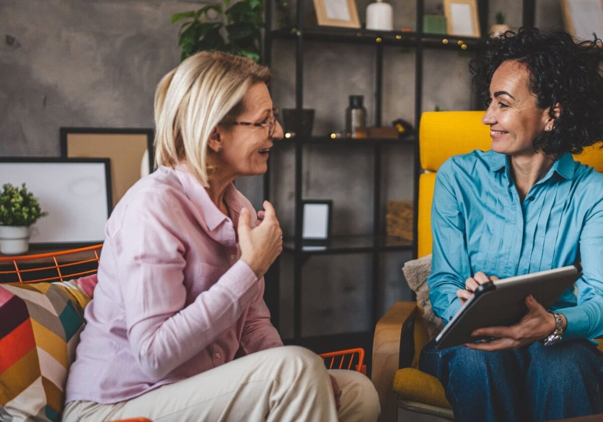 Two women smiling at each other while having a private conversation in comfortable chairs; one woman holds a tablet.