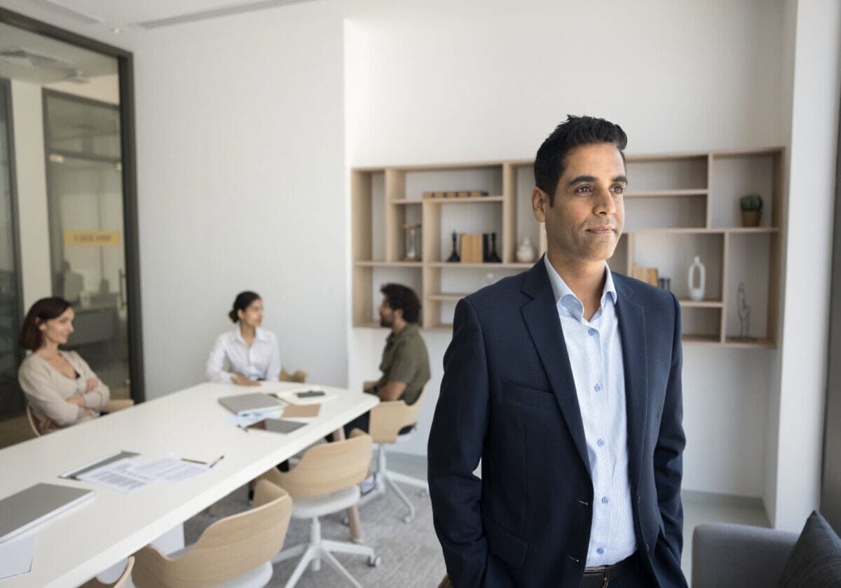 A confident man in a suit standing and looking out a window in a modern, bright office, with three colleagues seated at a conference table behind him.