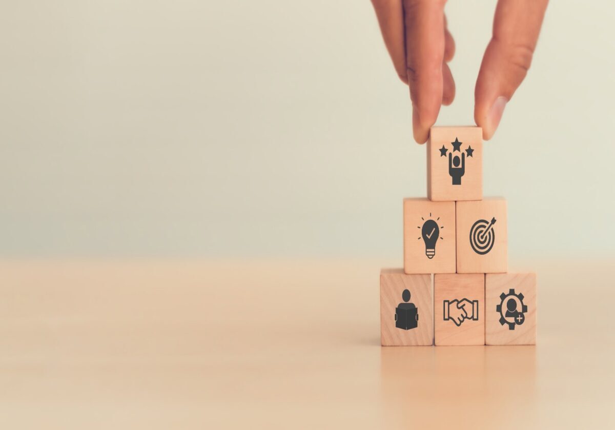 Hand placing a wooden block with three stars on top of a pyramid stack of blocks with business icons.