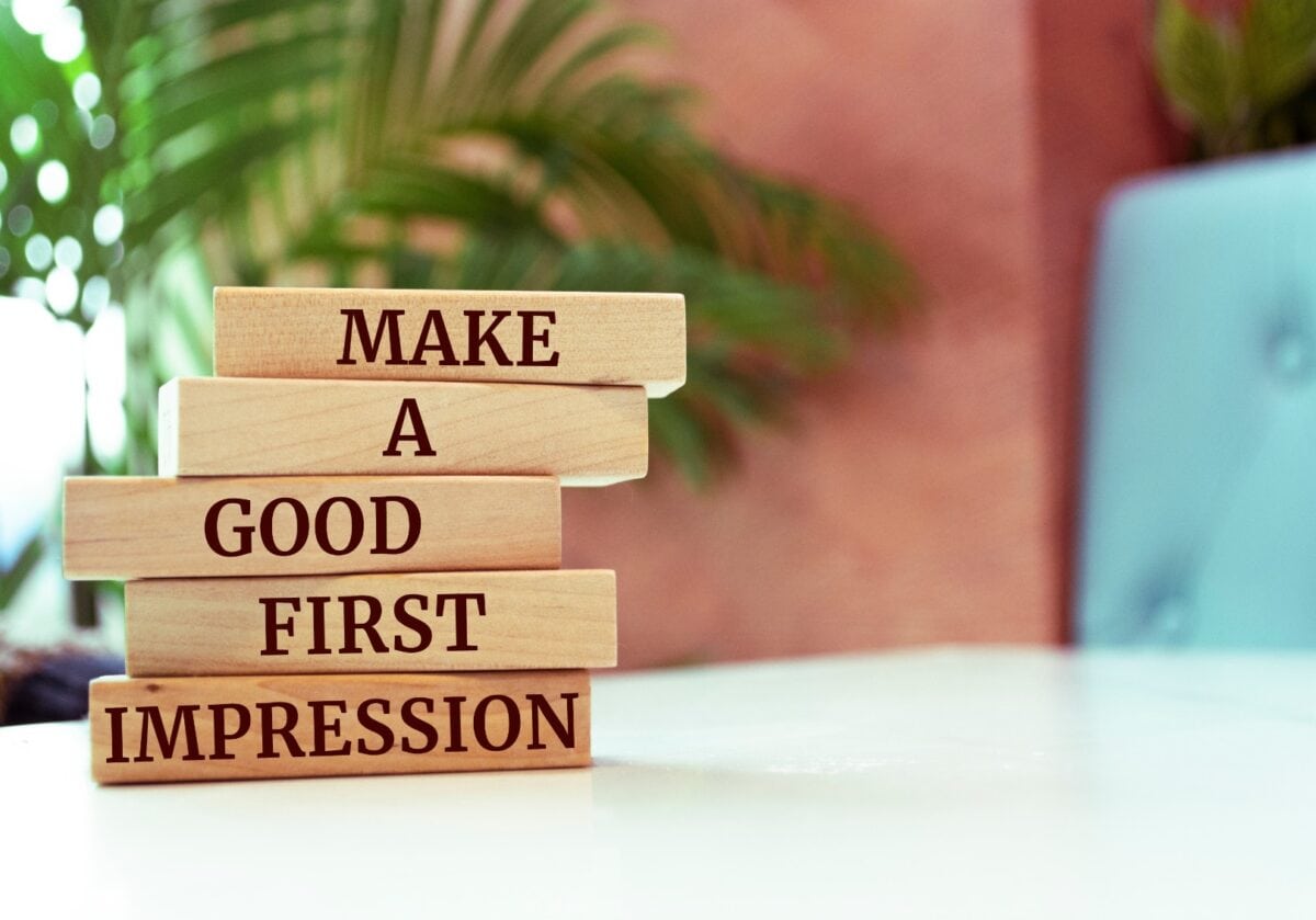 Wooden blocks stacked unevenly with the words “MAKE A GOOD FIRST IMPRESSION” printed on them, set against a blurred indoor background with plants.
