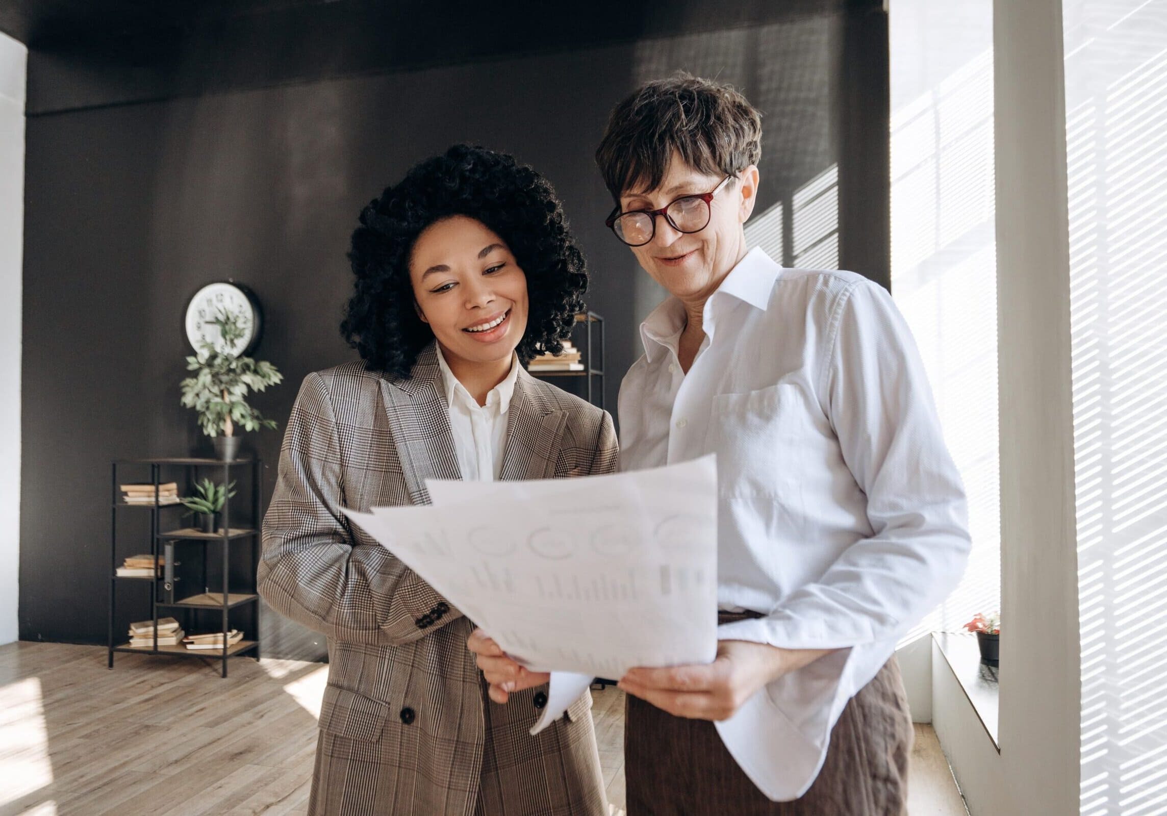 Two businesswomen smiling while reviewing printed documents with charts in an office