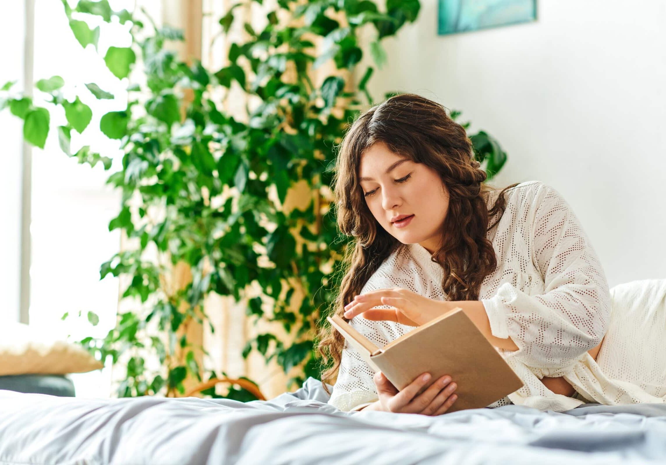 Young plus-size woman relaxing on a bed while reading a book in a bright, plant-filled bedroom