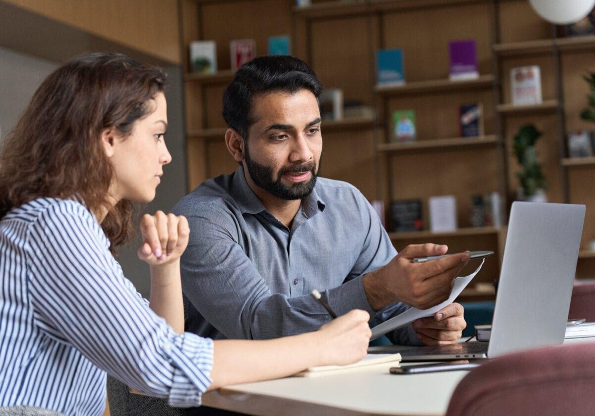 Two colleagues sit at a desk, discussing notes and looking at a laptop together in a modern office with bookshelves in the background.
