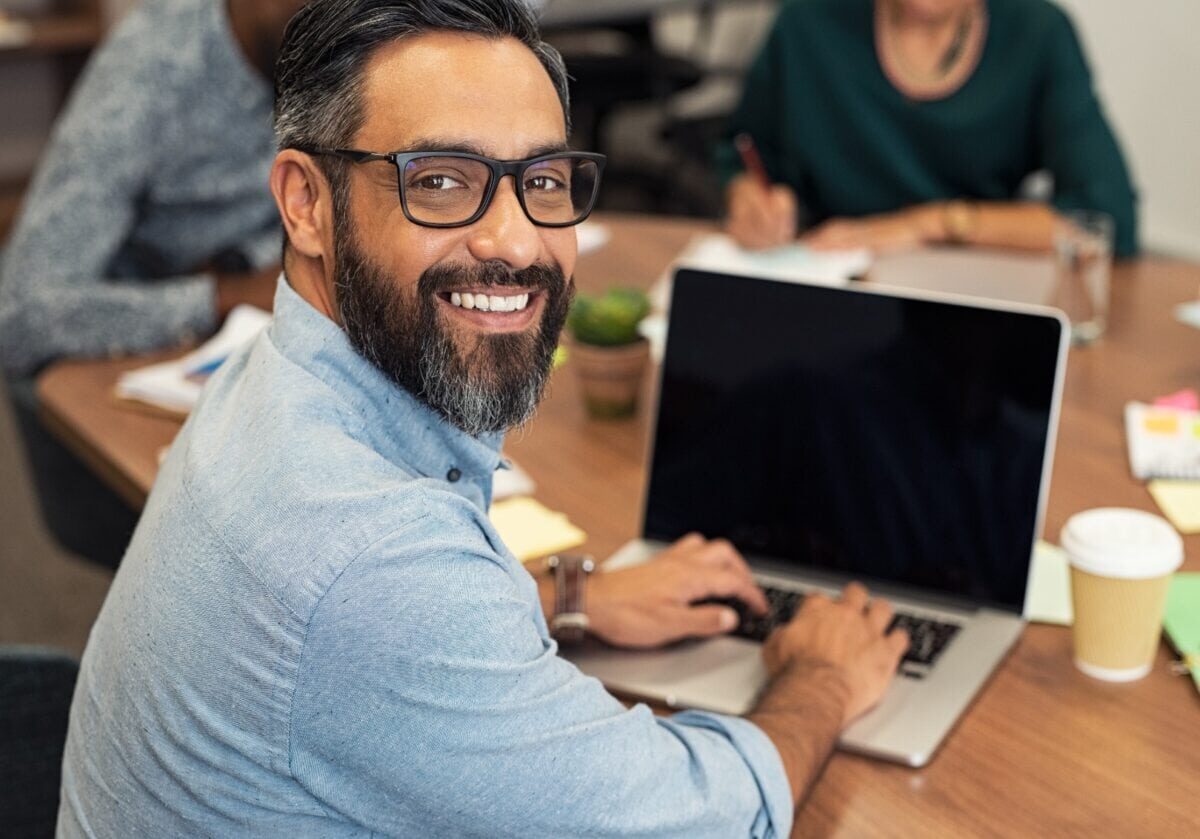 Smiling man with glasses looking over his shoulder while working on a laptop in a meeting.