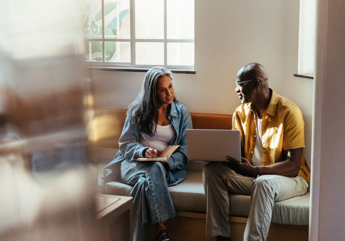 Female with long gray hair, wearing a denim jacket and white top, sits on a beige couch alongside a man in a yellow button-up shirt.