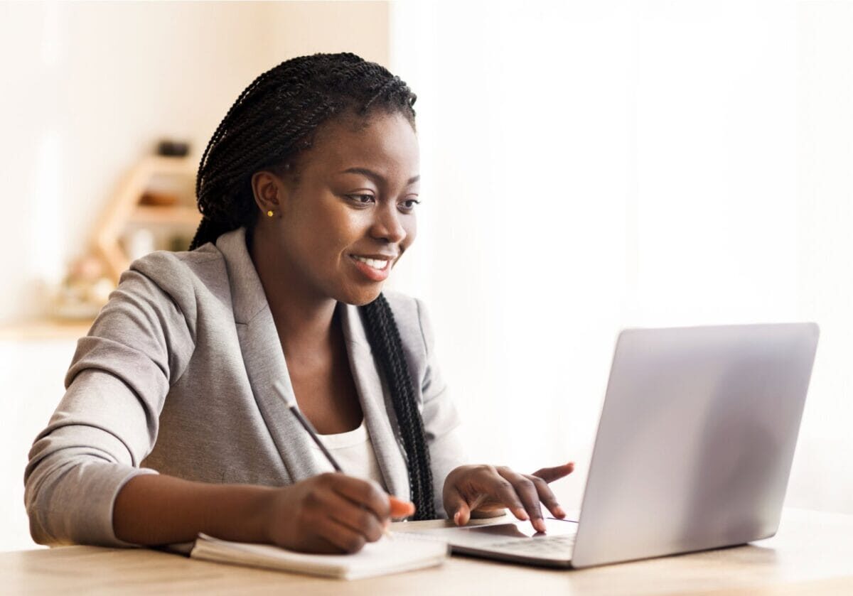 Smiling black professional writing in a notebook while looking at a laptop computer.