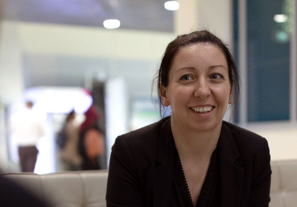 Jenn Header, woman with brown hair smiling wearing dark top sitting on a couch
