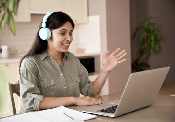 woman at a laptop with over ear headphones on smiling and waving in greeting while on video call with her coach