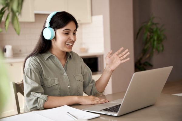 woman at a laptop with over ear headphones on smiling and waving in greeting while on video call with her coach