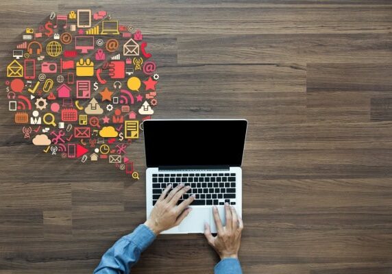 Person working at a computer with a large thought bubble to the left of the laptop comprised of many small icons typically associated with computers in varying colors