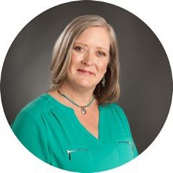Marilyn Orr PCC headshot photo, light haired smiling woman in a turquoise top in front of a gray background