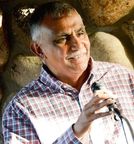 Ram Ramanathan ACTC MCC headshot photo, smiling man in button up shirt holding a microphone in front of a rock wall