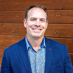 Richard Antosik ACC headshot photo, smiling man with short hair blue blazer and blue shirt standing in front of a wood wall