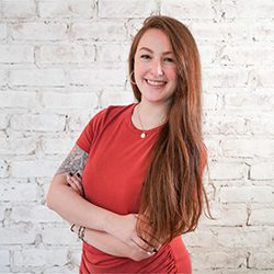 Sarah Orzach headshot photo, smiling red haired woman in a red top in front of a white brick wall