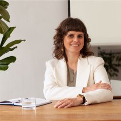 Lezanne Scott headshot photo, dark haired woman sitting at a table with a glass of water and a notebook smiling wearing a white blazer