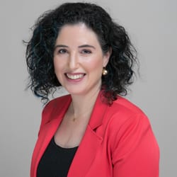 Shiri Ben-Arzi MCC headshot photo, smiling woman with dark hair wearing a bright red blazer and dark top in front of light gray background