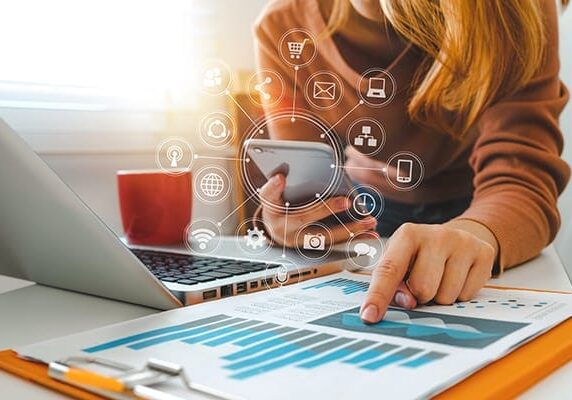 woman looking at data on a printed sheet and holding a cellphone with many connected dots and icons around it at a table with a laptop and coffee cup
