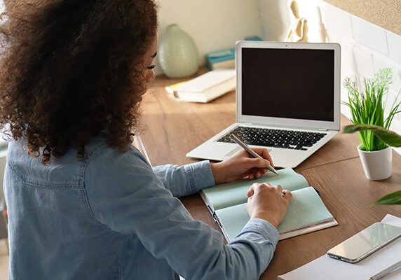 woman writing in a notebook at a desk with phone laptop plant and other papers