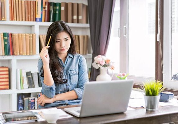 woman with pencil to her head looking stuck on a challenge sitting at a desk in a home office