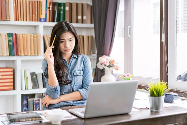 woman with pencil to her head looking stuck on a challenge sitting at a desk in a home office