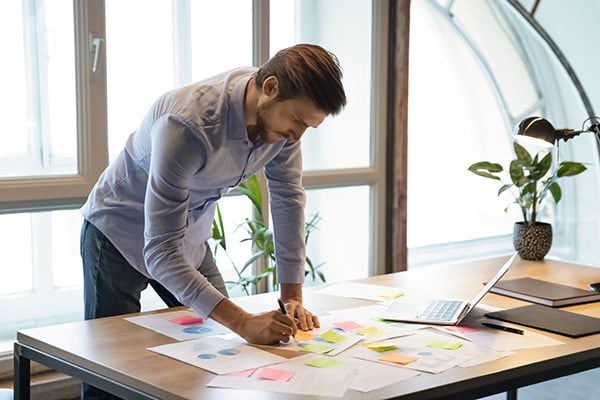 man working at a table with papers and sticky notes scattered as well as a laptop lamp and plant